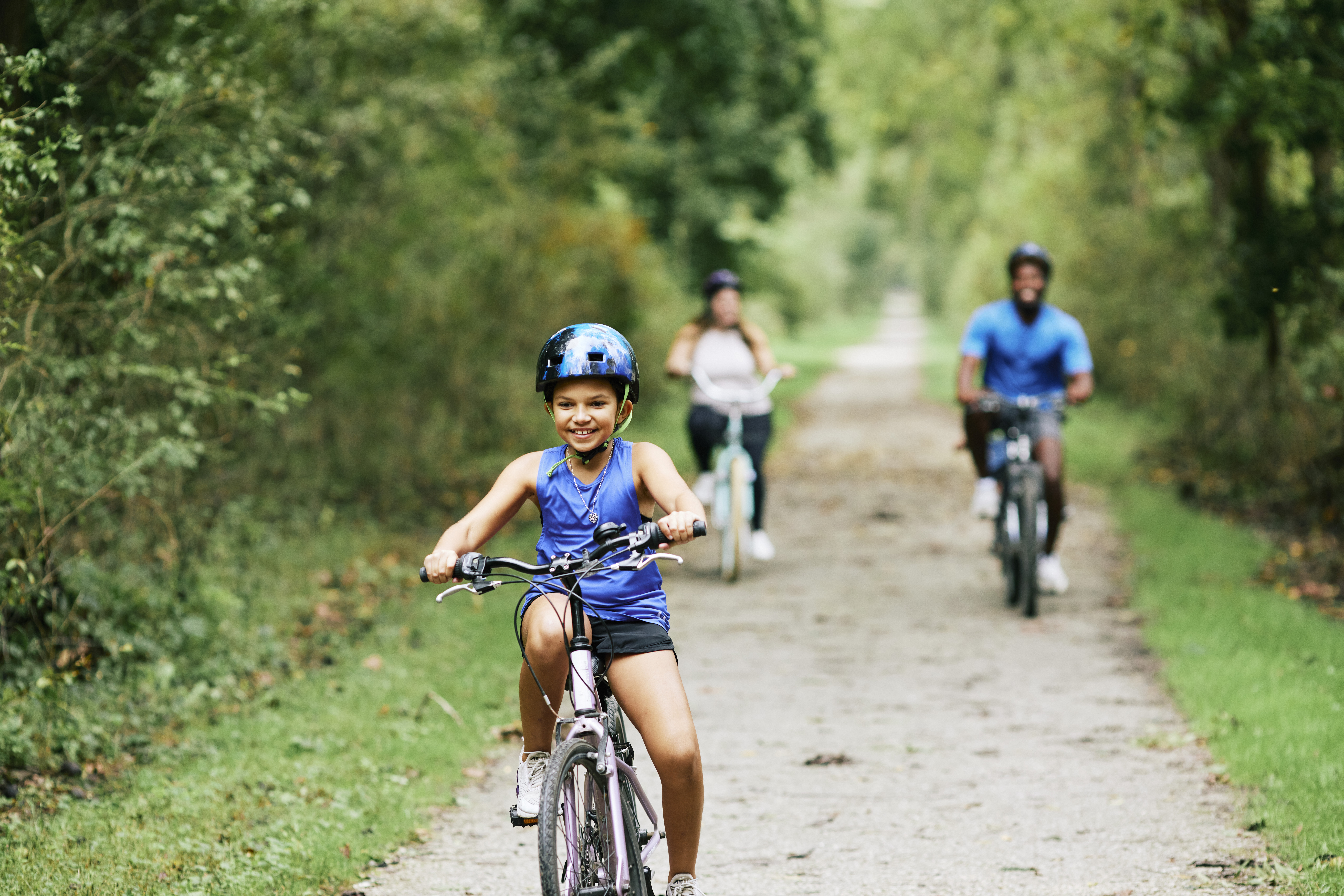 Little girl riding a bike on a wooded trail while smiling and with her parents riding bikes behind her.