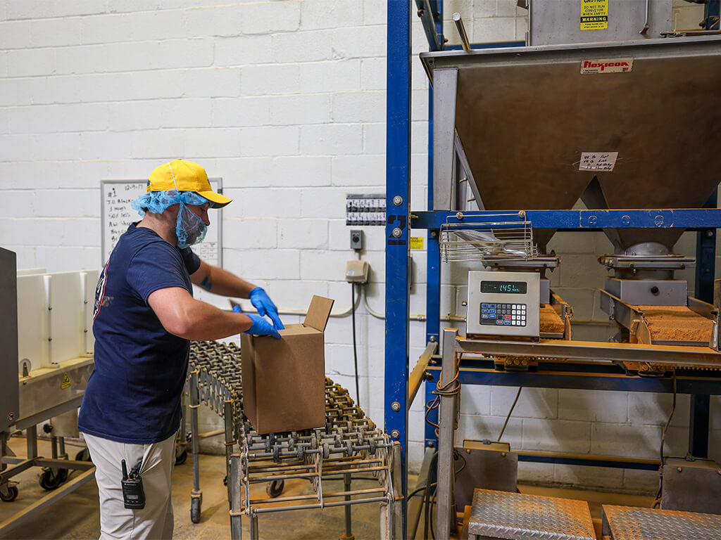 J.O. Spice Company employee packages crab seasoning at the Halethorpe warehouse, carefully filling a box beside industrial spice-blending equipment, reflecting the family-run operation’s hands-on production process.