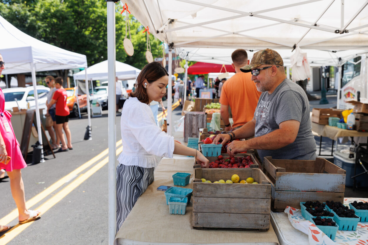 A customer picking fruit out of a wooden crate with a worker smiling at them under a tent at the Towson Farmers Market.