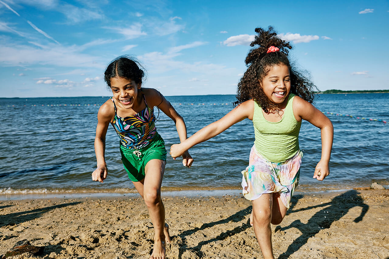 Two little girls holding hands and smiling while running away from the water at a beach in Baltimore County.
